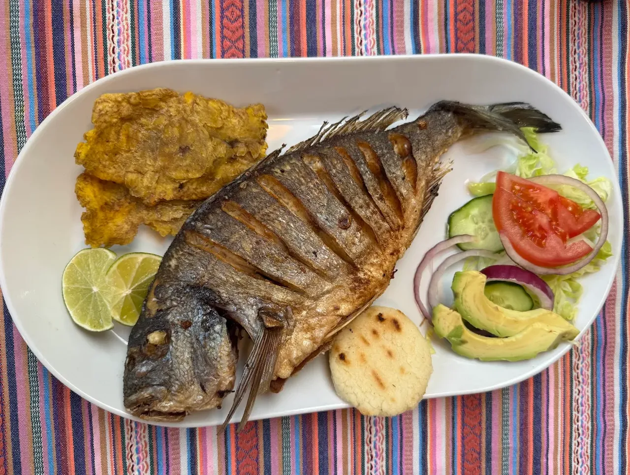 Hele gebakken dorade op een witte schaal met patacones, limoen, een arepa en salade van tomaat, komkommer, rode ui en avocado